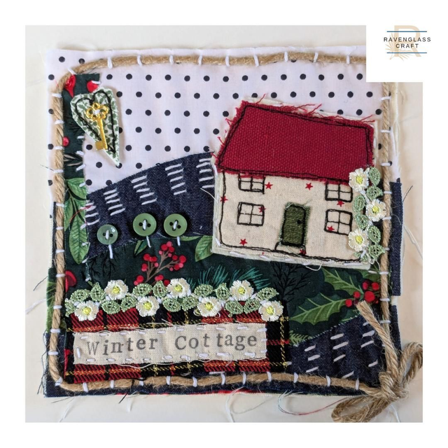 A square handmade fabric scene featuring an embroidered and appliqué winter cottage scene. The design includes a cream house with a red roof, dark navy hills, green foliage, white flowers, red berries, and a patch reading 'Winter Cottage.