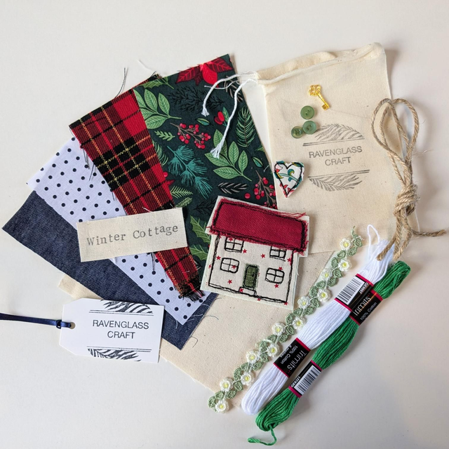A flat-lay of a craft kit featuring multiple fabric swatches (plaid, green leaf pattern, polka dots, navy), a small house patch, a cream canvas bag with 'RAVENGLASS CRAFT' branding, green and white embroidery floss, and a white paper tag.