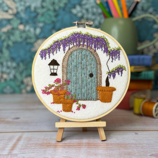 A circular embroidery hoop displaying a finished fantasy cottage embroidery design, mounted on a small wooden stand. The design features a light blue wooden door, purple wisteria, pink flowers, and potted plants on a white fabric background.