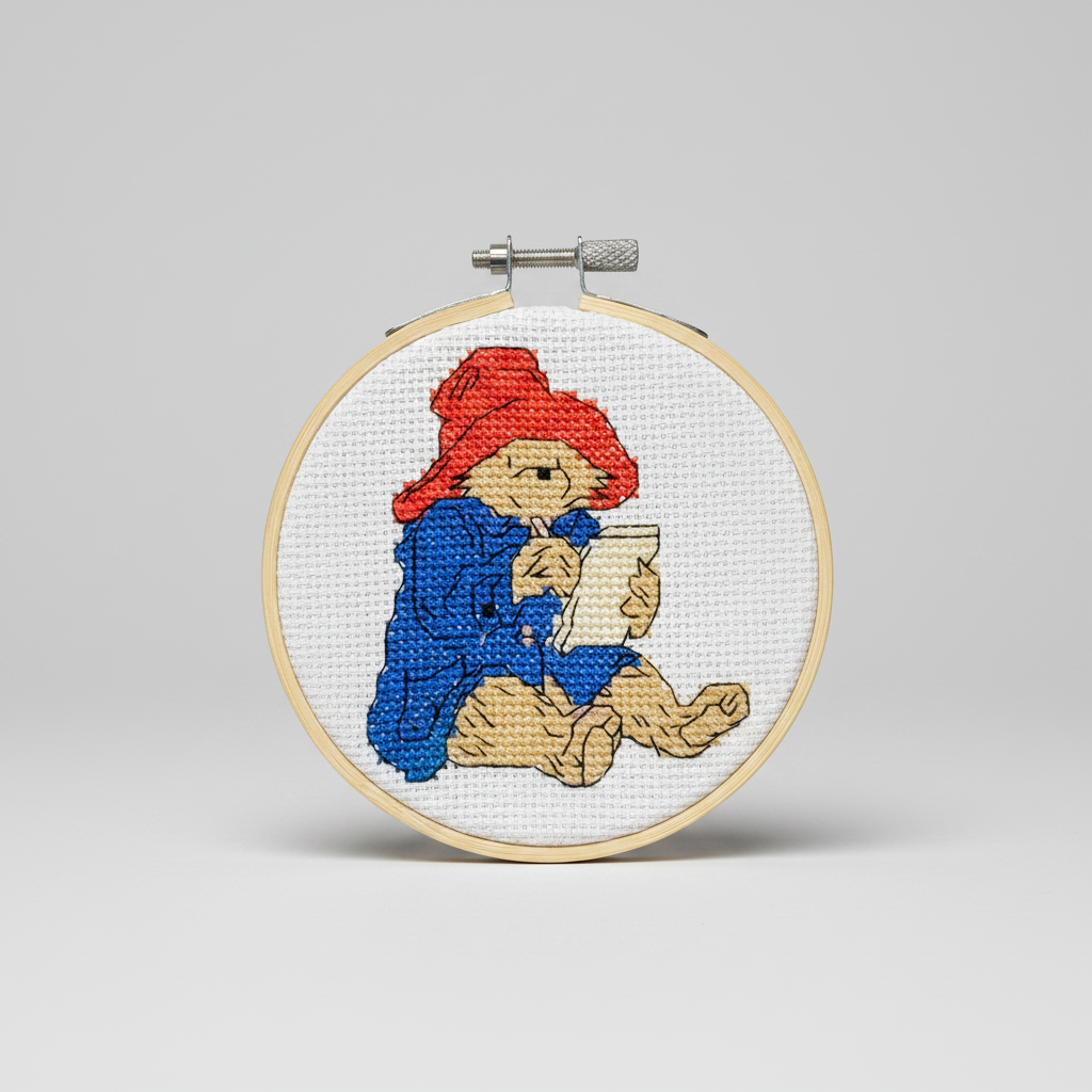 A circular embroidery hoop containing a cross-stitch design of Paddington Bear writing a letter. The bear is stitched in beige with a red hat and blue coat on a white fabric background.